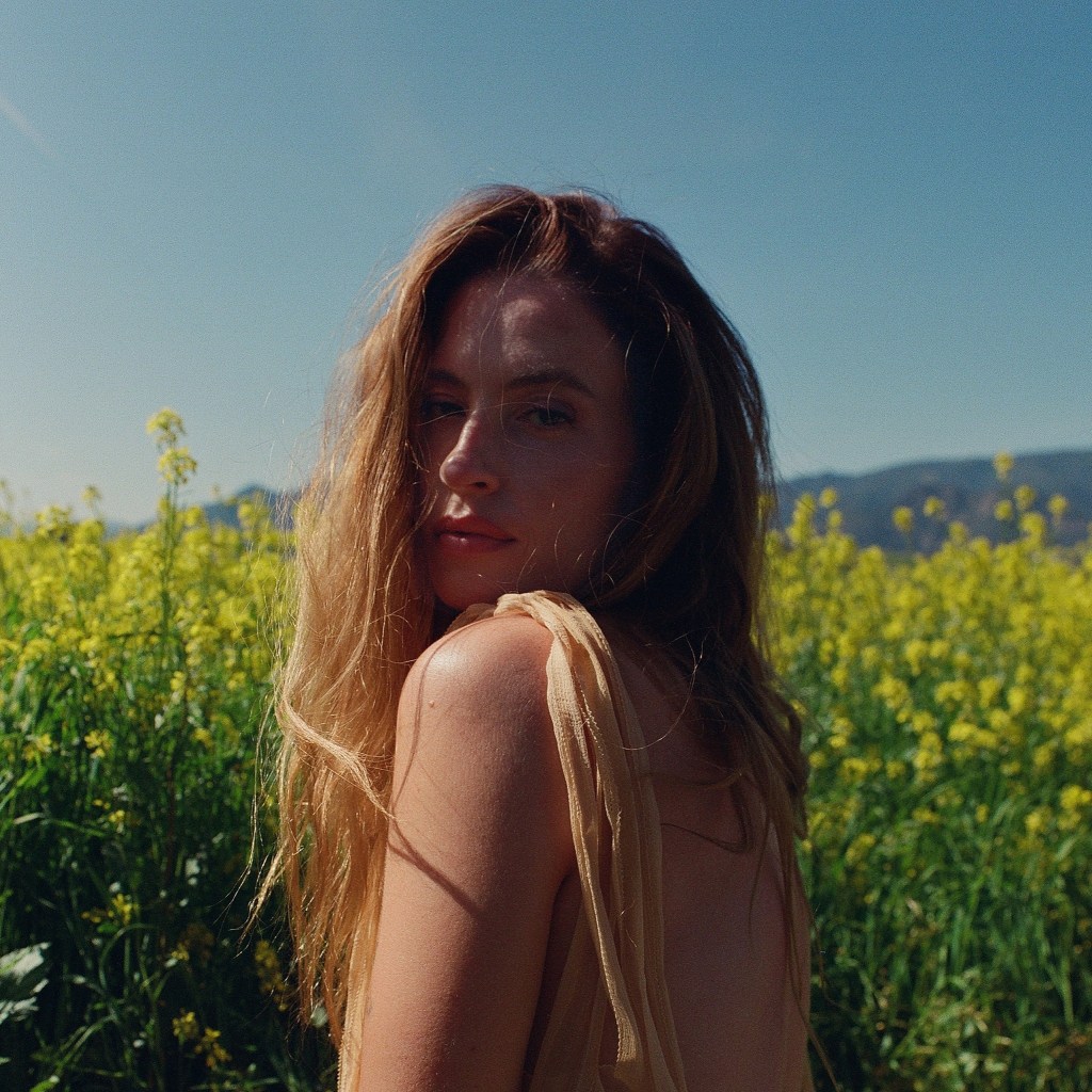 A photo from FLETCHER's Boy campaign. Fletcher is looking into the camera, wearing a beige sheer dress , in a field of yellow wildflowers.