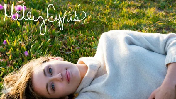 A photo of Miley Cyrus looking into the camera, wearing a beige knitted sweater on a grassy field.